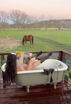 a woman sitting in a bathtub reading a book with a horse grazing in the background