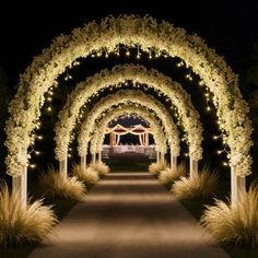 an archway decorated with white flowers and lit up at night in the background is a path that leads to a gazebo