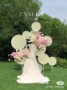 a woman in a wedding dress standing next to a large flower arrangement on top of a lush green field