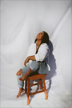 a woman sitting on top of a wooden chair next to a white wall with her eyes closed