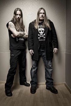 two men with long hair standing next to each other in front of a white wall