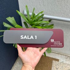 a person holding up a red and gray sign that says sala 1 in front of a plant