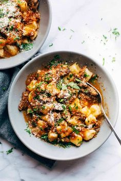 two bowls filled with pasta and meat on top of a white tablecloth next to silver spoons