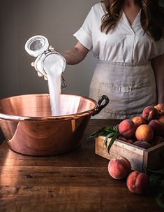 a woman pouring milk into a copper bowl on top of a wooden table next to fruit