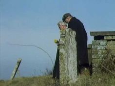 two people standing next to each other on top of a grass covered hill near a stone wall