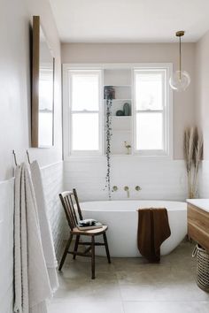 a white bath tub sitting next to a wooden chair in a bathroom under two windows