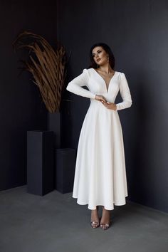 a woman standing in front of a plant wearing a white dress with long sleeves and deep v - neck