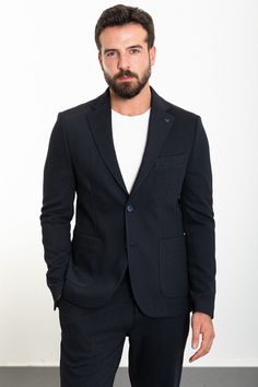 a man with a beard wearing a black suit and white t - shirt standing in front of a wall