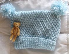 a teddy bear laying on top of a blue knitted hat with pom poms