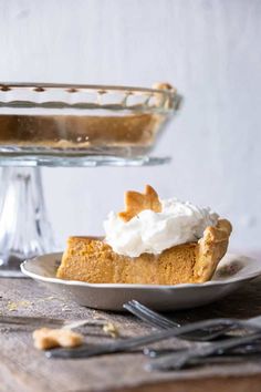 a slice of pumpkin pie on a plate with whipped cream in the foreground and a glass cake stand behind it