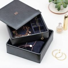 two black boxes with jewelry on top of a white table next to a plant and perfume bottle