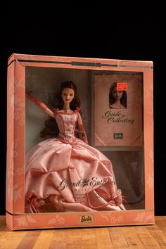 a barbie doll in a box on a wooden table with a black background and pink dress