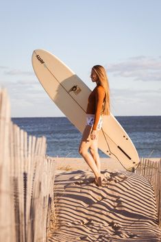 a beautiful young woman holding a surfboard on top of a sandy beach next to the ocean