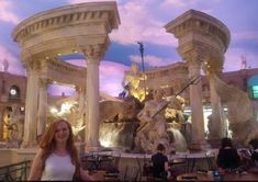 a woman standing in front of a fountain with statues on it and people sitting at tables