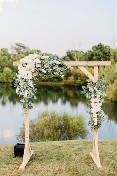 a wedding arch with flowers and greenery on the grass near a lake in front of a wooden structure