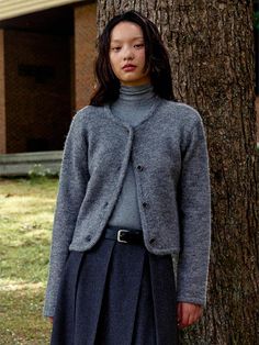 a woman standing in front of a tree wearing a gray sweater and pleated skirt