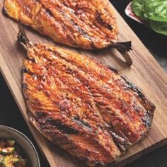 two steaks on a wooden cutting board next to a bowl of vegetables and sauce