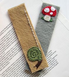 two bookmarks made out of felt sitting on top of an open book, one has a green leaf and the other is a red mushroom