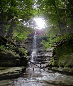 two people standing in front of a waterfall with trees on either side and sunlight coming through the falls