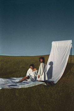 two women sitting on top of a white sheet in a grassy field next to each other