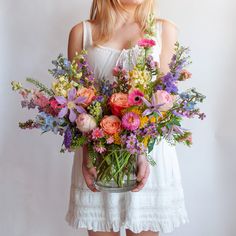 a woman in white dress holding a vase filled with colorful flowers and greenery on her chest