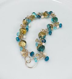 This beautiful bracelet features Czech glass beads, wrapped in 14k Gold Filled wire. The main bracelet consists of Deep Teal Blue beads with a Gold patina and Aurora Borealis luster. I added Deep Ocean Blue, Ivory AB, Aqua, and Clear Gold-Lined beads to create a full bracelet. Everything has been meticulously hand wrapped with 14k Gold Filled wire. The hook & eye clasp is hand forged by me, as always! One of the things I love about cluster bracelets is their beautiful movement. This one is no ex Gold Hand-strung Czech Glass Bracelets, Gold Czech Glass Hand-strung Bracelet, Hand-strung Gold Czech Glass Jewelry, Hand-strung Gold Jewelry With Czech Glass, Gold Czech Glass Jewelry, Hand-strung, Gold Glass Bracelets With Faceted Beads, Gold Bracelets With Faceted Beads, Wire Wrapped Czech Glass Bracelets With Round Beads, Unique Gold Beaded Glass Bracelets