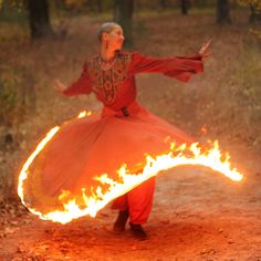 a woman in an orange dress is dancing with fire on a dirt road surrounded by trees