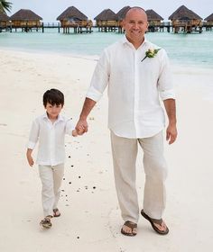 an older man holding the hand of a young boy on a beach with thatched huts in the background