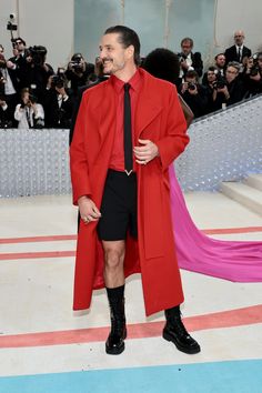 a man in a red coat and black tie standing on a blue carpet with cameras around him