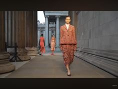 two models walk down the catwalk in front of columns and pillars, wearing orange dresses