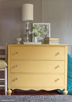 a yellow dresser sitting next to a white chair in a living room with a lamp on top of it