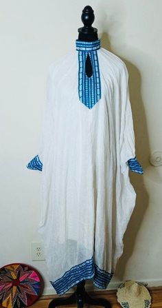 Ethiopian and Eritrean beautiful simple traditional  dress or cultural piece with blue and white patterns. It has a loose fit with decorative blue embroidery or trim at the neck, cuffs, and hem. Traditional White Thobe With Patterns, Traditional Blue Cotton Kaftan, White Long Sleeve Kurta With Traditional Patterns, Traditional White Kaftan For Spring, Traditional White Thobe With Long Sleeves, Traditional White Long Sleeve Thobe, White Long Sleeve Kaftan For Traditional Ceremonies, White Long Sleeve Thobe For The Beach, Traditional White Tunic Kaftan