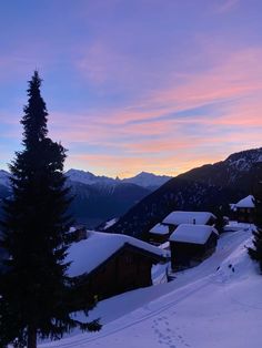 the sun is setting over some houses on a snowy mountain side with trees and mountains in the background