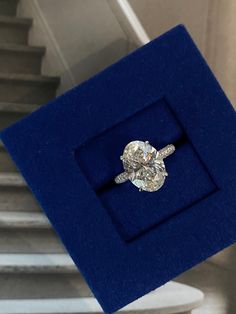 an oval diamond ring sits in a blue velvet box on top of some white stairs