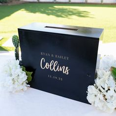 a black box with gold lettering on it and white flowers in front of the box