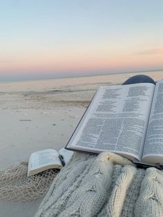 an open book sitting on top of a blanket next to the ocean with a sunset in the background