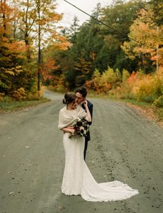 a bride and groom standing in the middle of a dirt road surrounded by fall foliage