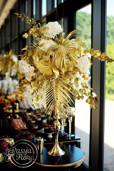 a gold vase filled with lots of flowers on top of a black table next to a window