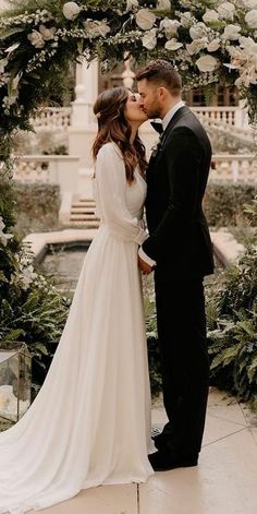 a bride and groom kissing in front of an arch with white flowers on the side