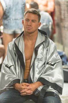 a man sitting on top of a stool wearing a silver jacket and black pants with his shirt open