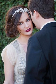 a man and woman standing next to each other in front of some bushes with their heads together