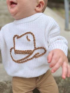 a smiling baby wearing a sweater with a hat on it's chest and brown pants