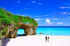 two people are walking on the beach near an arch in the sand and blue water