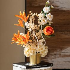 an arrangement of flowers in a gold vase on top of a stack of books next to a wooden wall