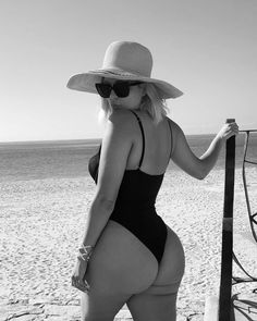 a woman in a bathing suit and hat standing on the beach next to a fence