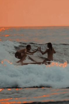 two people on surfboards in the ocean with waves coming towards them and an orange sky