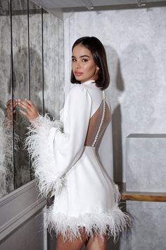 a woman wearing a white dress with feathers on the skirt and heels standing in front of a mirror