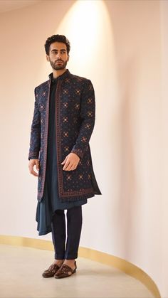 a man standing in front of a white wall wearing a black and blue jacket with an intricate pattern