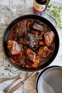 a pan filled with meat and vegetables on top of a table next to a bottle of wine