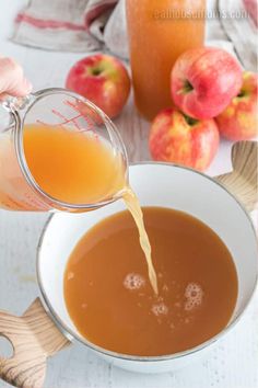 someone pouring caramel apple cider into a white bowl filled with orange juice and apples in the background